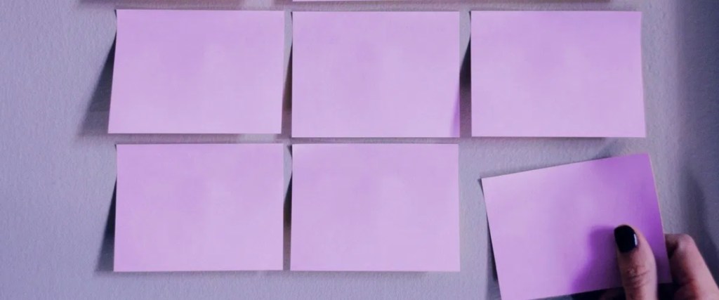 Metodologías ágiles en Nu Colombia Una mano pega una nota adhesiva morada clarita en una pared de color similar.