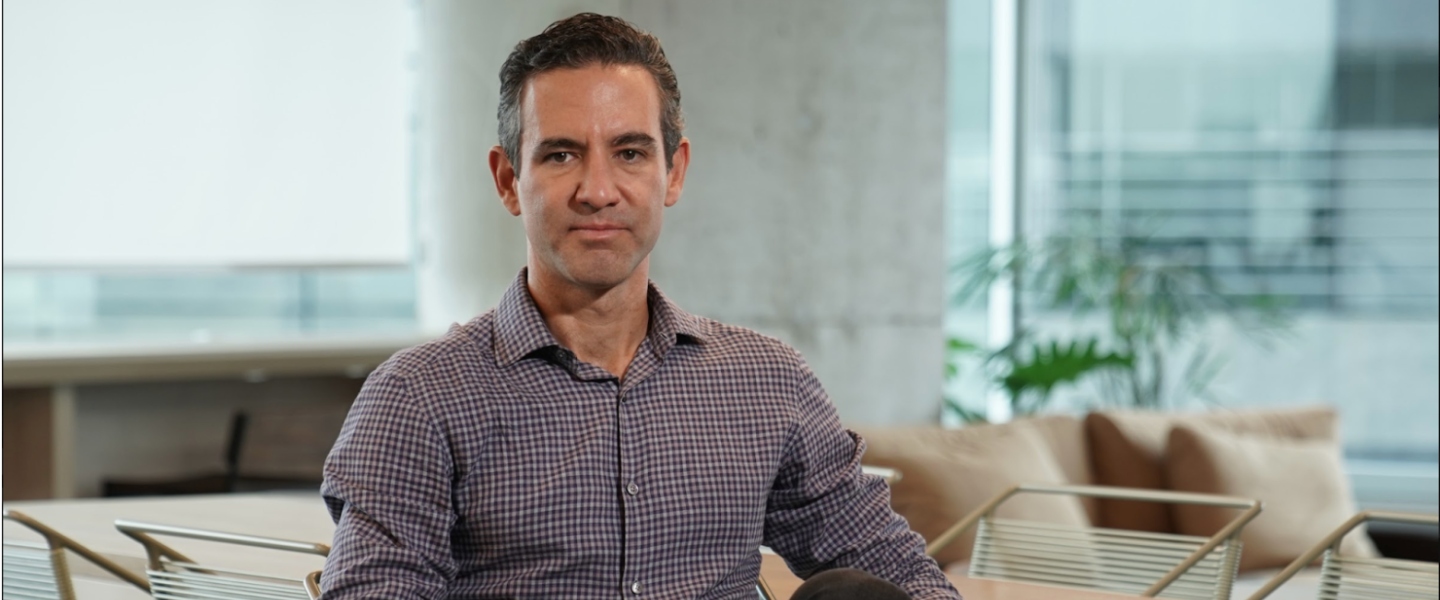 David Vélez: La historia de Nu David Vélez posa sentado en una oficina con una camisa a cuadros y jeans negros.