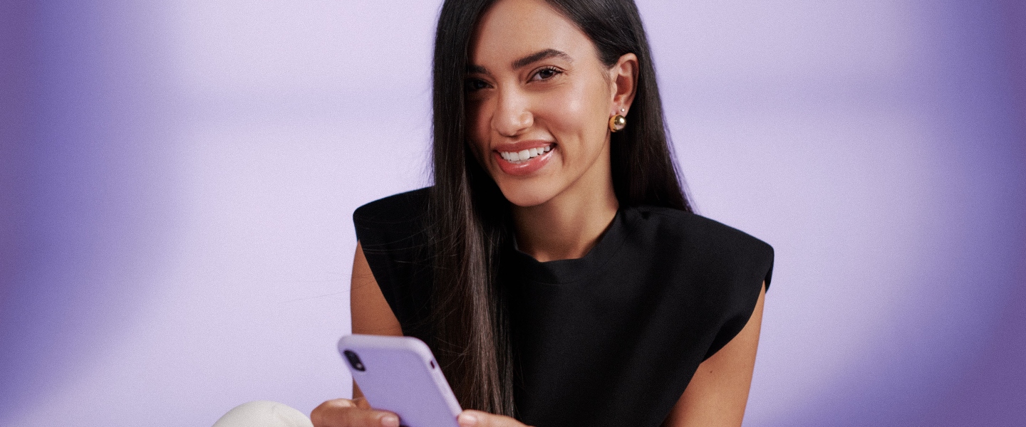 Una mujer joven, morocha, sonríe mientras sostiene un celular de color lila