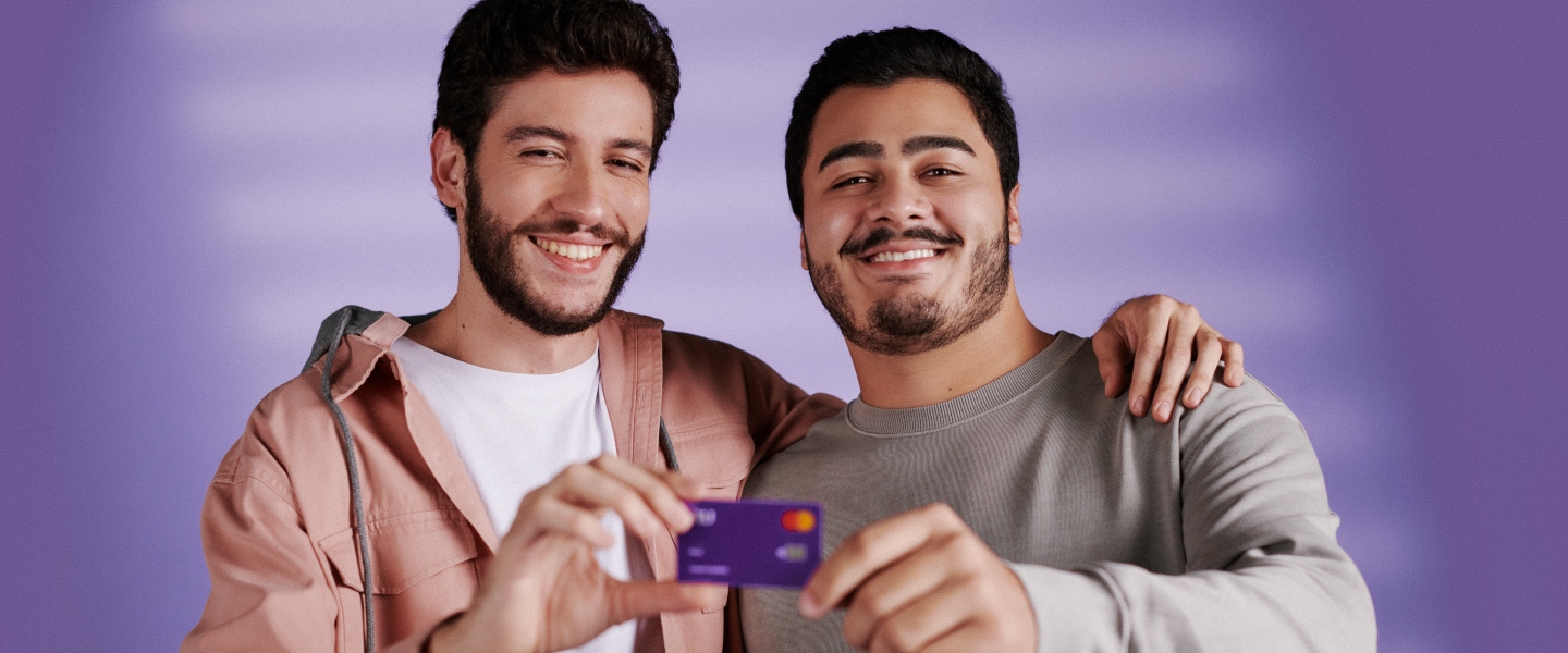 Dos hombres jóvenes se abrazan y posan sosteniendo ambos con sus manos una tarjeta de crédito Nu, la Moradita