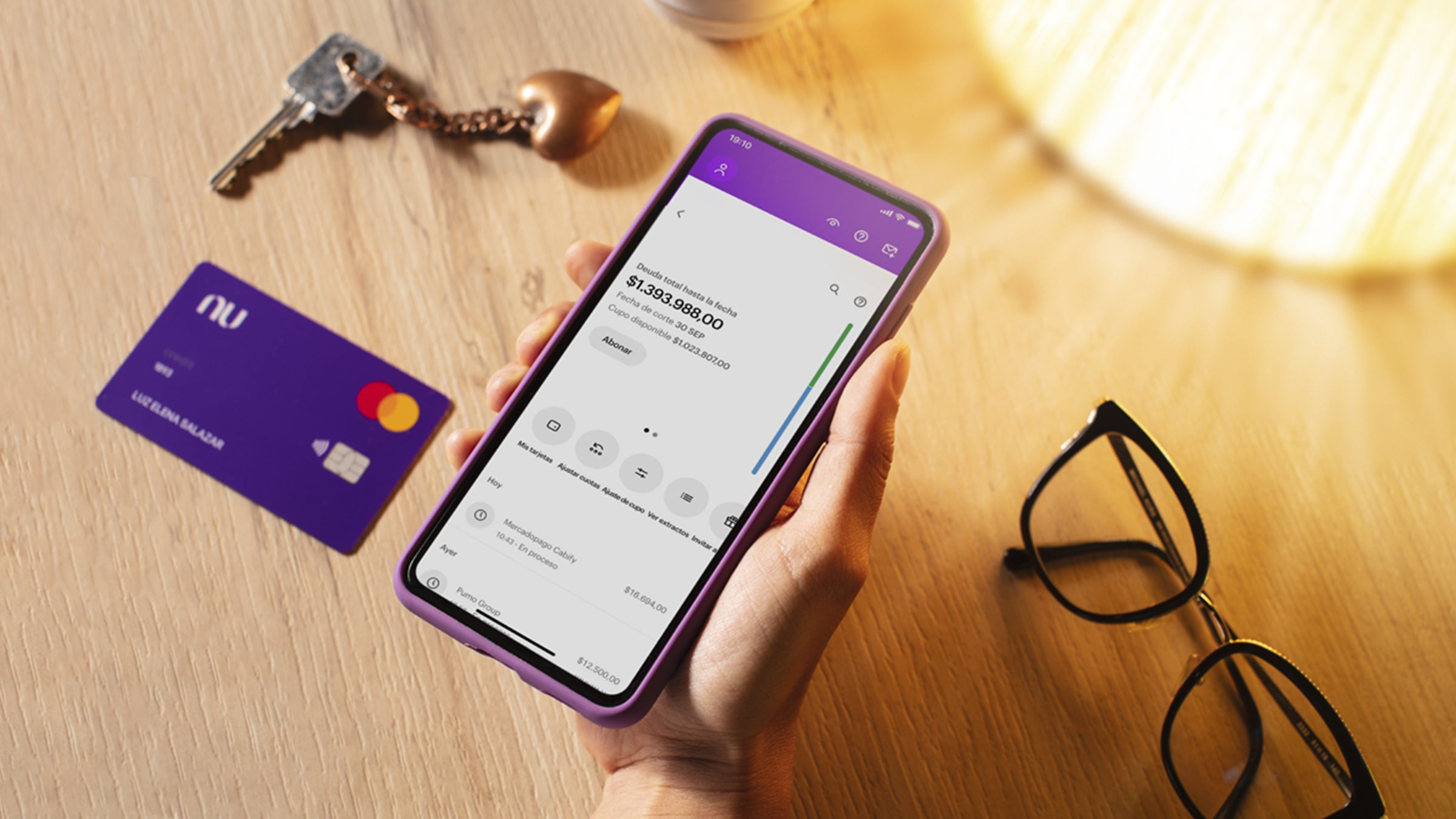 App Nu: Controla tus finanzas desde la palma de tu mano Foto de una mano con el celular y el app de Nu abierta, en el fondo la tarjeta de Nu y unas llaves